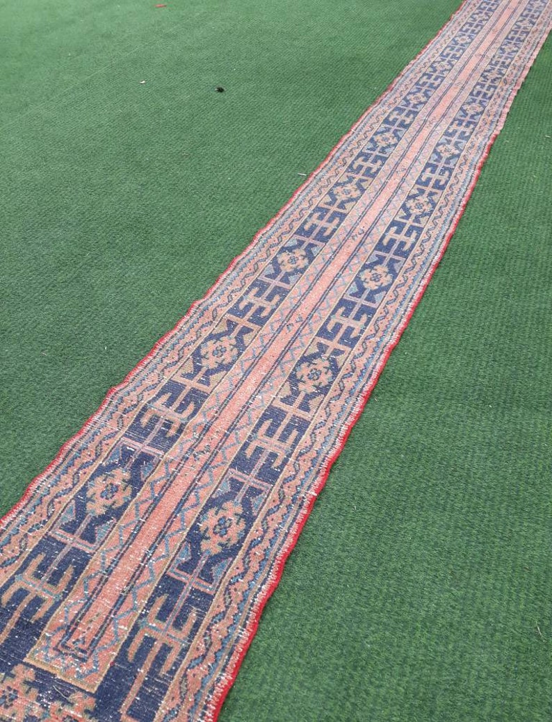 May include: A long, narrow rug with a pink, blue, and white geometric pattern. The rug is laid out on a green surface.