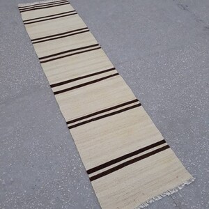 May include: A long, narrow, cream-coloured rug with brown stripes. The rug has a fringe on both ends.