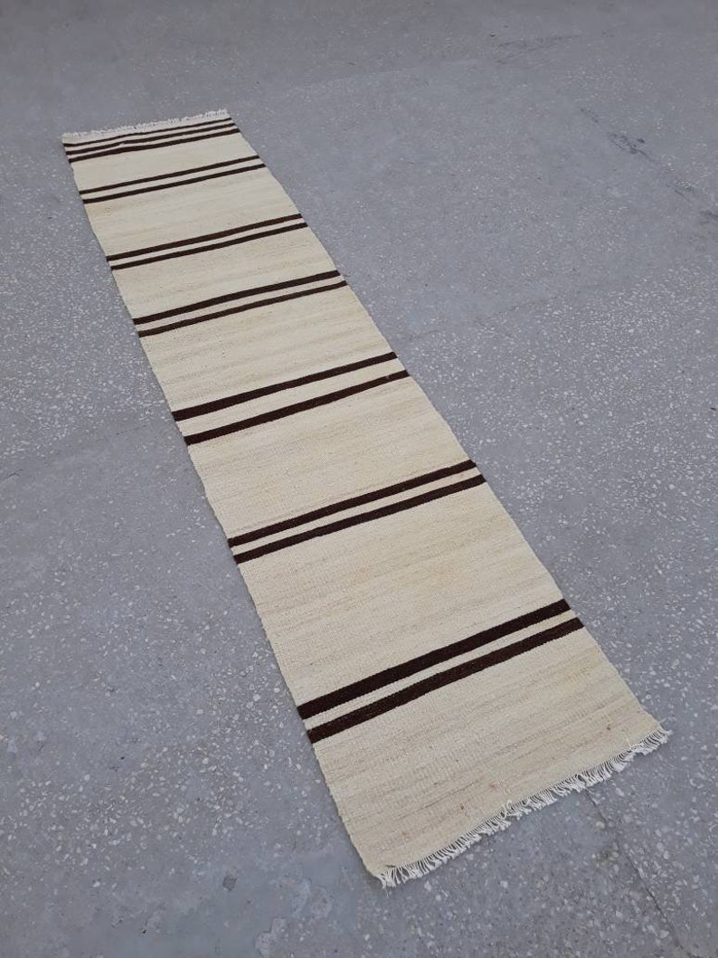 May include: A long, narrow, cream-coloured rug with brown stripes. The rug has a fringe on both ends.