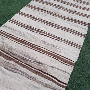 May include: A white and brown striped rug with a fringed edge. The rug is laid out on a green artificial grass surface.