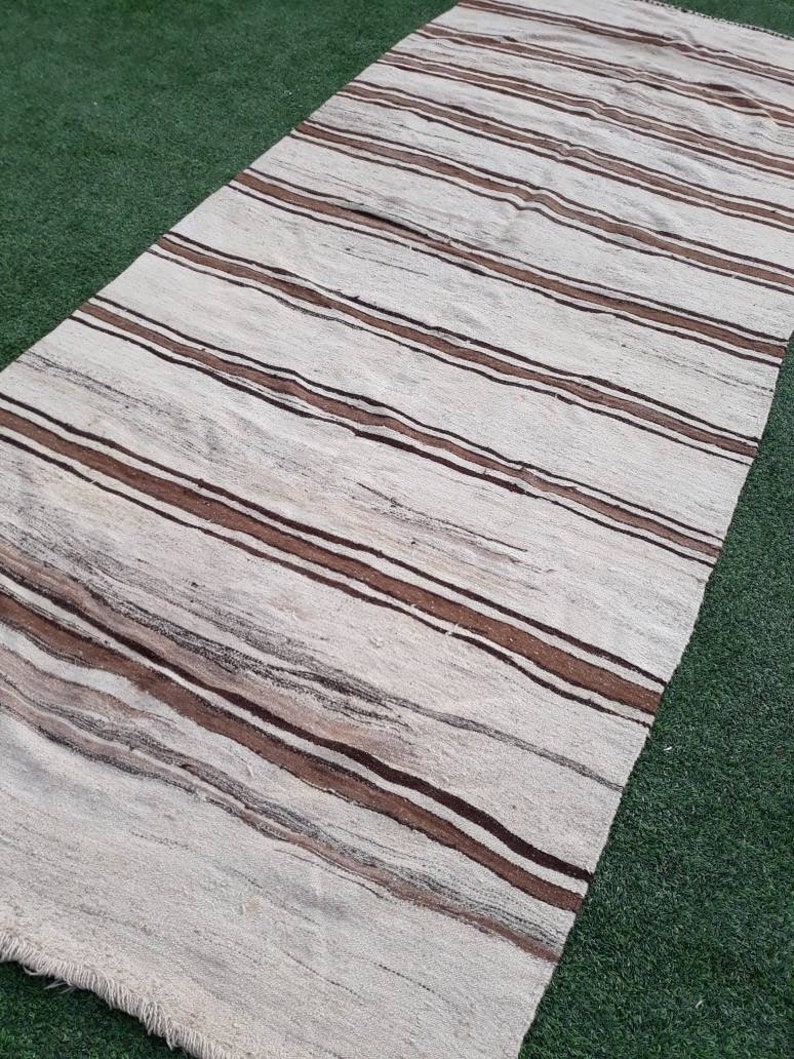 May include: A white and brown striped rug with a fringed edge. The rug is laid out on a green artificial grass surface.