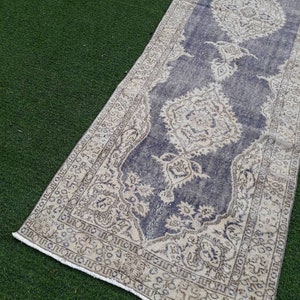 May include: A vintage runner rug with a faded blue background and a cream-colored floral pattern. The rug has a frayed edge and is laid out on a green artificial grass surface.