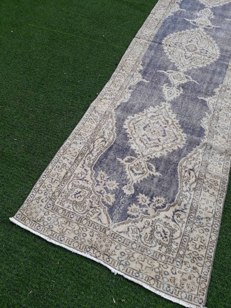 May include: A vintage runner rug with a faded blue background and a cream-colored floral pattern. The rug has a frayed edge and is laid out on a green artificial grass surface.