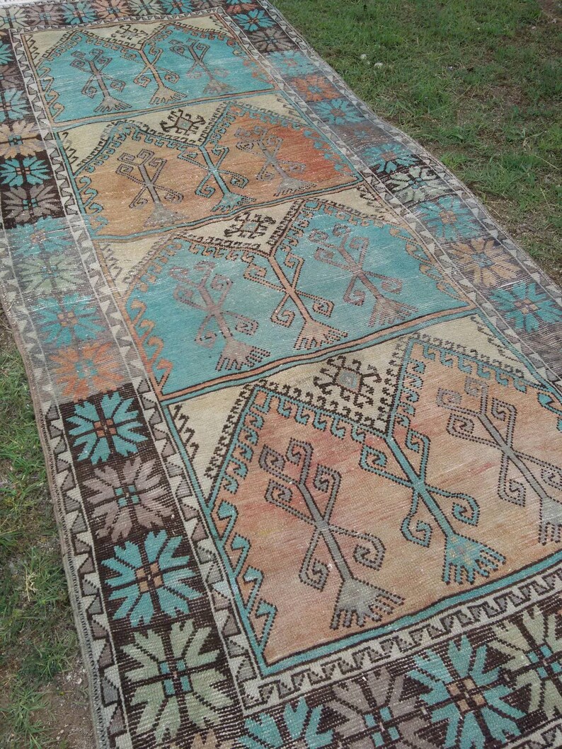 Puede incluir: Una alfombra turca vintage de corredor con un esquema de color turquesa, marr&oacute;n y crema. La alfombra presenta un patr&oacute;n geom&eacute;trico con motivos florales estilizados.
