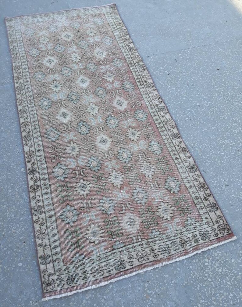 K&ouml;nnte beinhalten: Ein Vintage-L&auml;ufer mit einem floralen Muster in Rosa-, Beige- und Gr&uuml;nt&ouml;nen. Der Teppich hat einen verblassten, abgenutzten Look und einen Fransenrand.