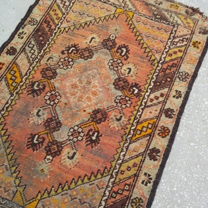 May include: A vintage Turkish rug with a faded pink, brown, and yellow geometric pattern. The rug has a fringed edge and is made of wool.