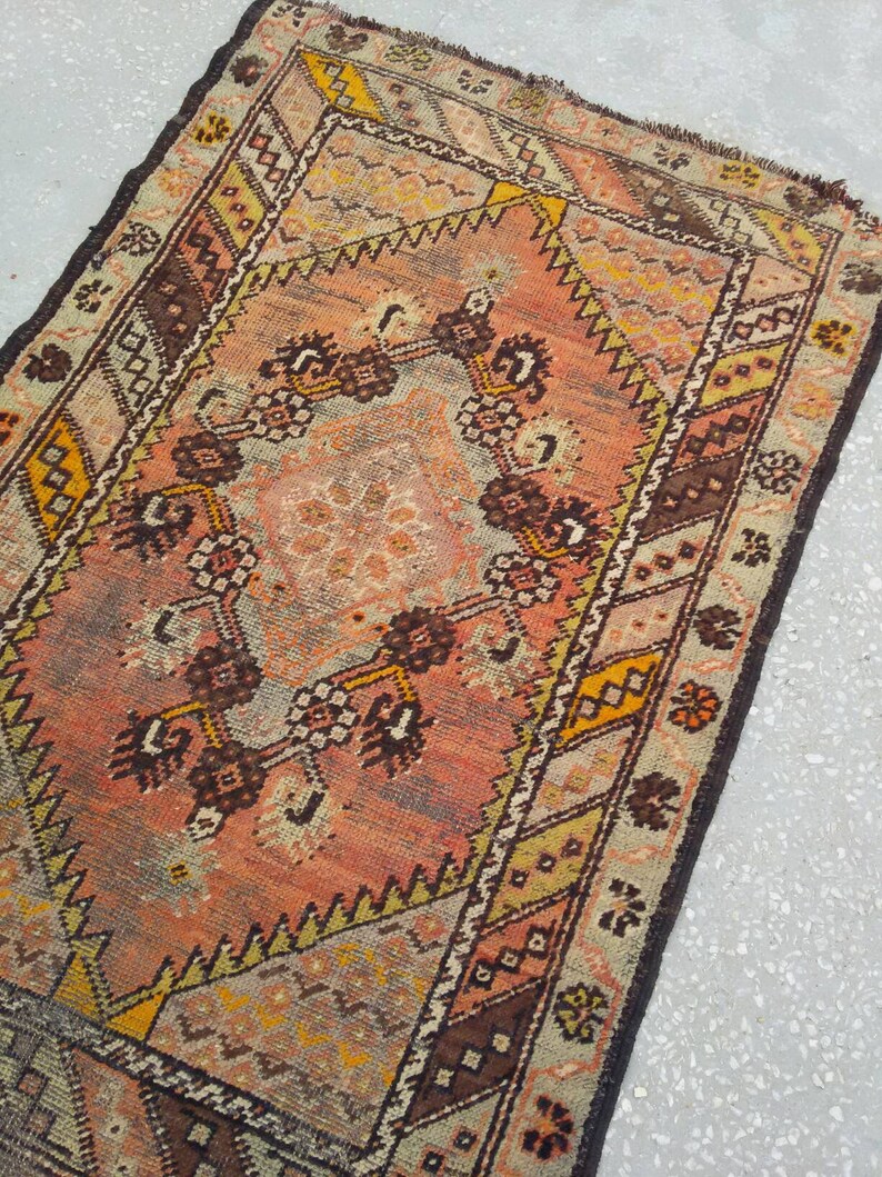 May include: A vintage Turkish rug with a faded pink, brown, and yellow geometric pattern. The rug has a fringed edge and is made of wool.