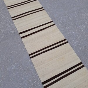 May include: A long, narrow, off-white rug with brown stripes. The rug has a fringe on the bottom edge.