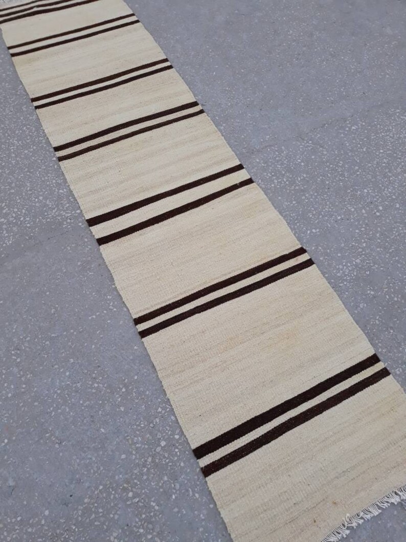 May include: A long, narrow, off-white rug with brown stripes. The rug has a fringe on the bottom edge.