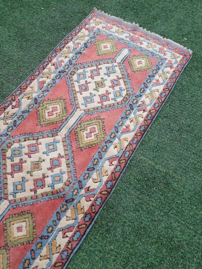 May include: A red, blue, and green patterned rug with a geometric design. The rug has a fringed edge and is laid out on a green surface.