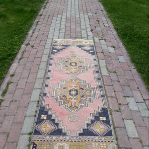 May include: A pink and yellow patterned runner rug with a dark blue border. The rug is laid out on a brick walkway.