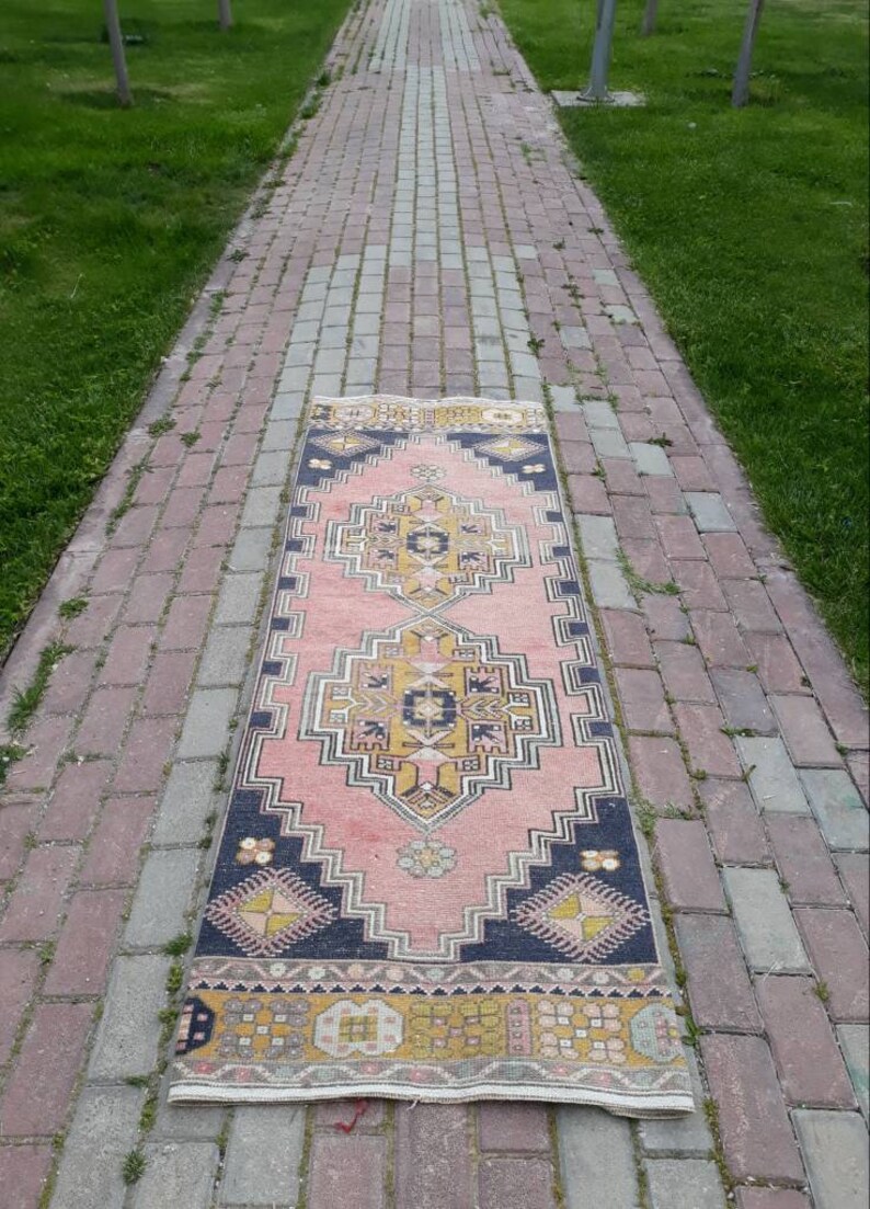 May include: A pink and yellow patterned runner rug with a dark blue border. The rug is laid out on a brick walkway.