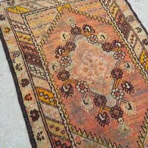 May include: A vintage Turkish rug with a faded pink, brown, and yellow geometric pattern. The rug has a worn and distressed look, with a fringed edge.