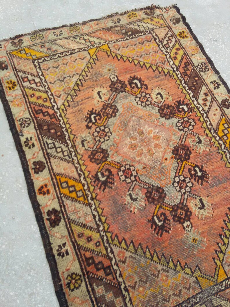 May include: A vintage Turkish rug with a faded pink, brown, and yellow geometric pattern. The rug has a worn and distressed look, with a fringed edge.