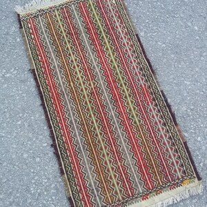 May include: A rectangular rug with a geometric pattern in shades of red, green, brown, and white. The rug has a fringe border.