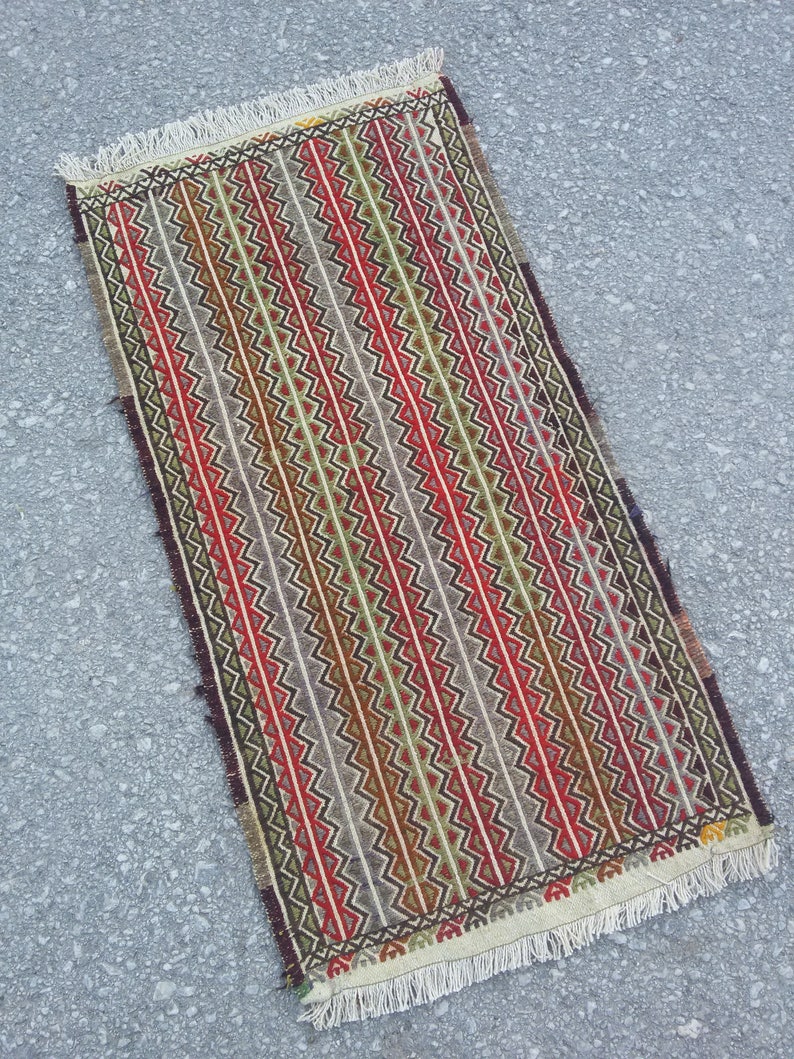 May include: A rectangular rug with a geometric pattern in shades of red, green, brown, and white. The rug has a fringe border.