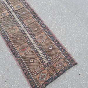 May include: A long, narrow rug with a traditional Persian design. The rug is in shades of brown, pink, and cream, with a central stripe of white. The rug has a fringed edge.