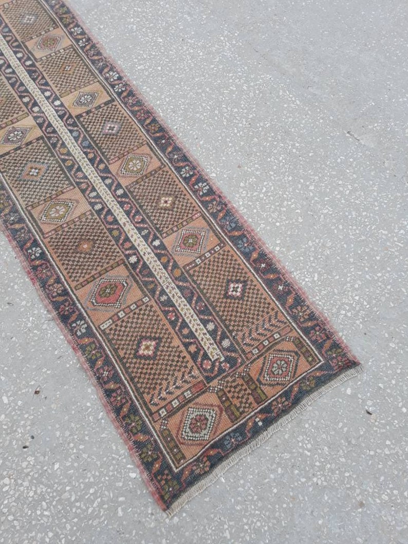 May include: A long, narrow rug with a traditional Persian design. The rug is in shades of brown, pink, and cream, with a central stripe of white. The rug has a fringed edge.