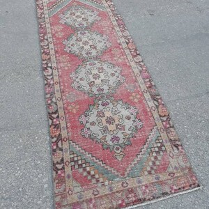 May include: A vintage Turkish runner rug with a pink background and intricate floral patterns in shades of beige, gray, and pink. The rug has a fringed edge and is approximately 2 feet wide and 6 feet long.