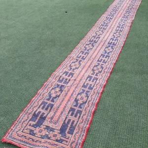 May include: A long, narrow rug with a traditional Persian design. The rug has a red background with blue and white geometric patterns. The rug is lying on a green artificial grass surface.