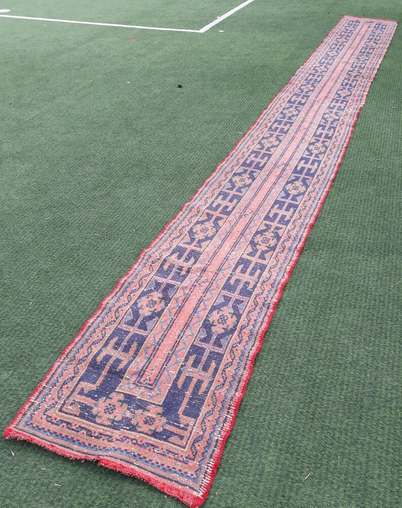 May include: A long, narrow rug with a traditional Persian design. The rug has a red background with blue and white geometric patterns. The rug is lying on a green artificial grass surface.