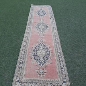 May include: A long, pink runner rug with a traditional Persian design. The rug has a dark blue and cream border with a repeating floral pattern. The center of the rug features three large, intricate medallions.