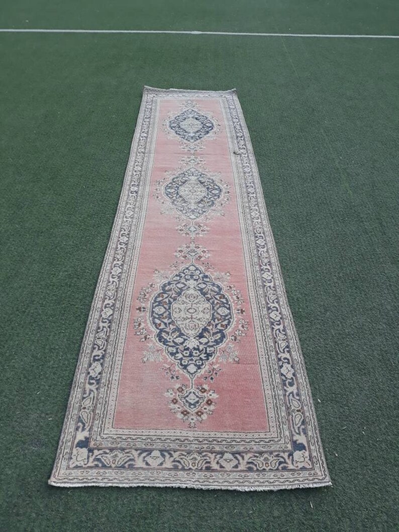 May include: A long, pink runner rug with a traditional Persian design. The rug has a dark blue and cream border with a repeating floral pattern. The center of the rug features three large, intricate medallions.
