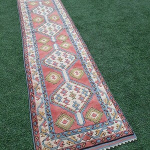 May include: A long, narrow rug with a geometric pattern in shades of pink, blue, green, and white. The rug has a fringed edge.