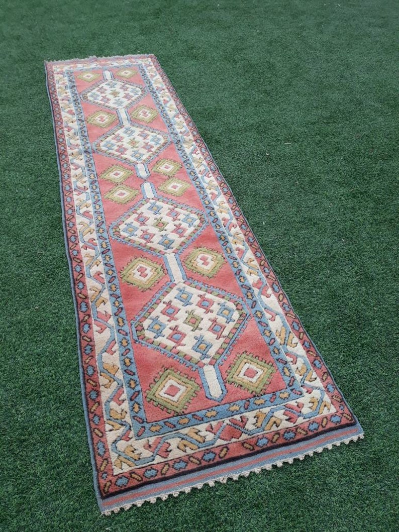 May include: A long, narrow rug with a geometric pattern in shades of pink, blue, green, and white. The rug has a fringed edge.