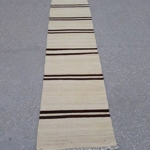 May include: A long, narrow, off-white rug with brown stripes. The rug has a fringe on the bottom edge.