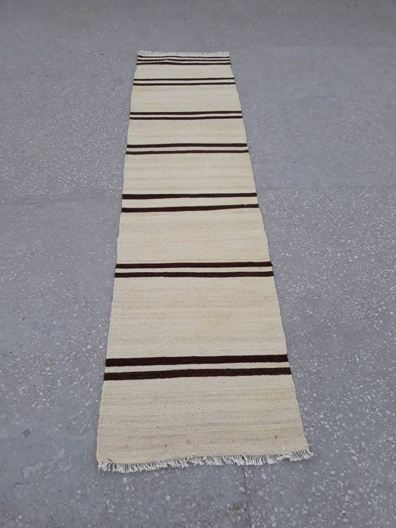 May include: A long, narrow, off-white rug with brown stripes. The rug has a fringe on the bottom edge.