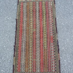 May include: A traditional handwoven rug with a geometric pattern in shades of red, green, brown, and white. The rug has a fringe border.
