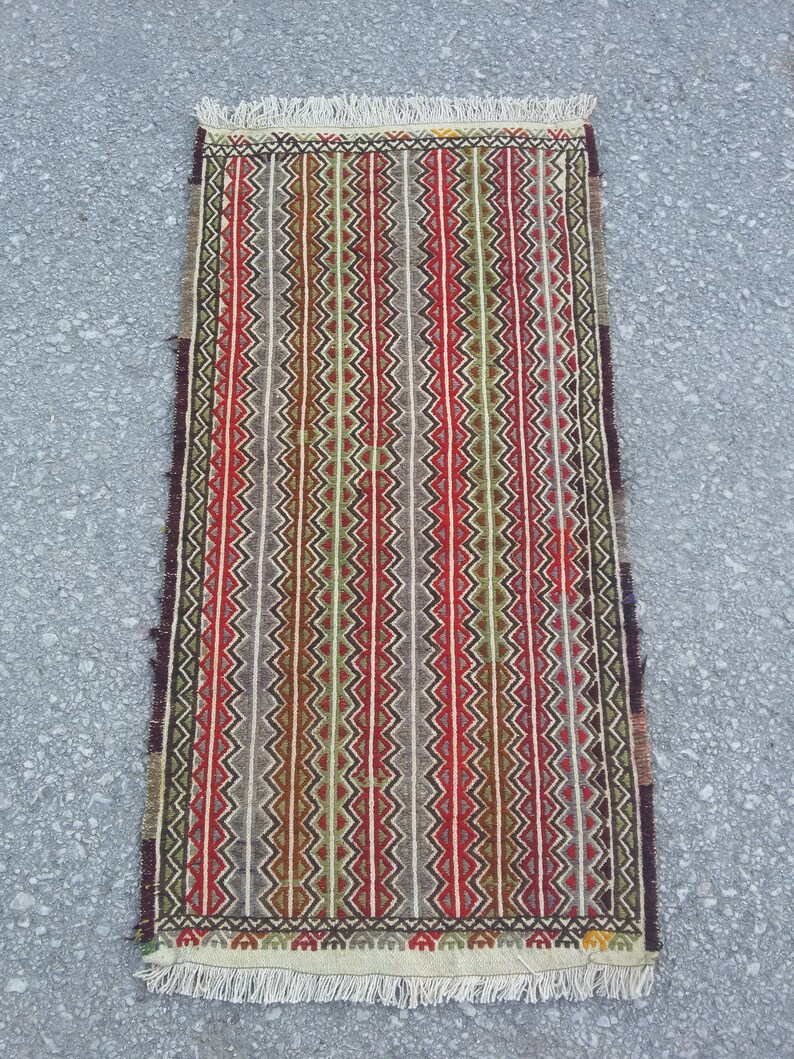 May include: A traditional handwoven rug with a geometric pattern in shades of red, green, brown, and white. The rug has a fringe border.