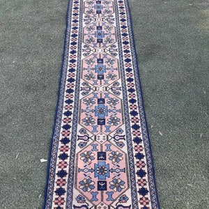 May include: A long, narrow rug with a pink, blue, and white geometric pattern. The rug has a dark blue border with a repeating floral design.