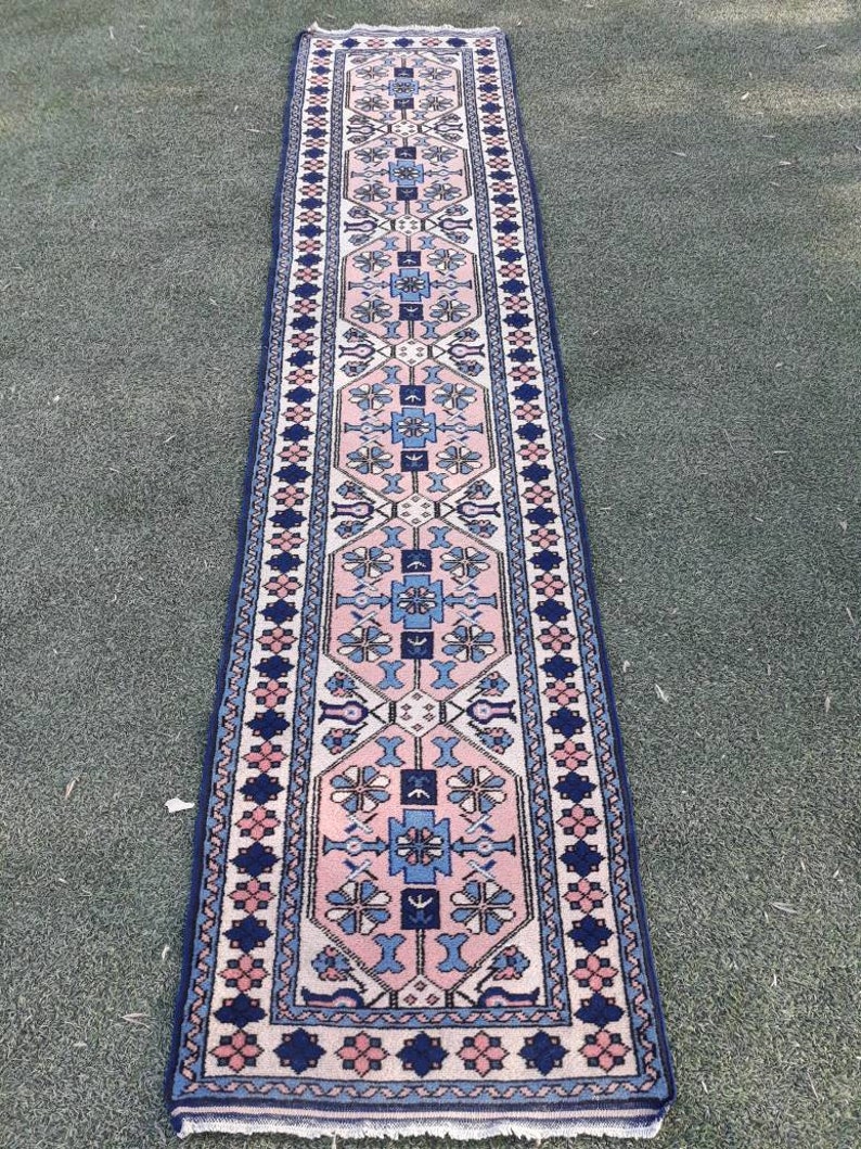 May include: A long, narrow rug with a pink, blue, and white geometric pattern. The rug has a dark blue border with a repeating floral design.