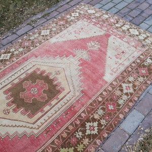 May include: A vintage Turkish rug with a red, brown, and cream color scheme. The rug has a geometric pattern with a central diamond shape.