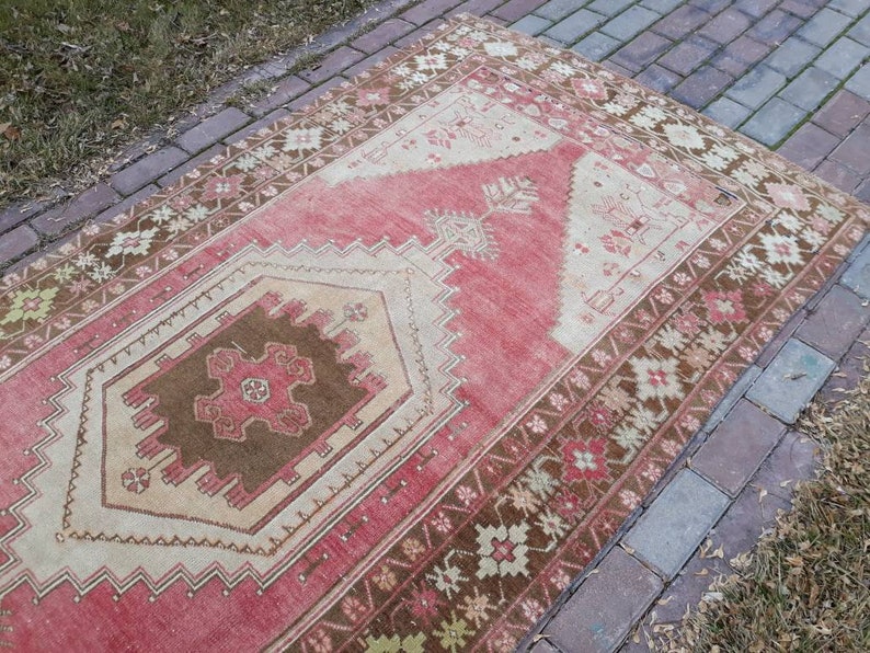 May include: A vintage Turkish rug with a red, brown, and cream color scheme. The rug has a geometric pattern with a central diamond shape.