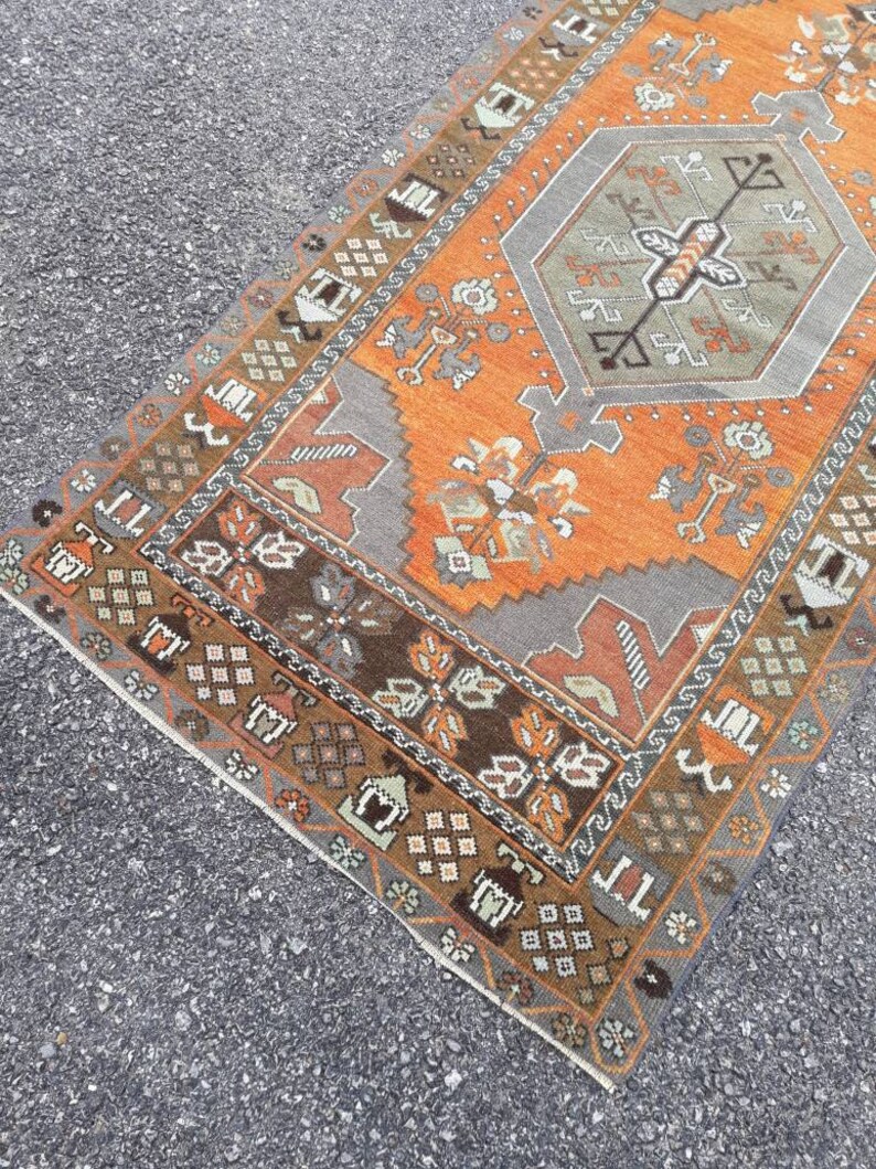 May include: A vintage Turkish rug with a geometric pattern in shades of orange, brown, gray, and white. The rug has a fringed edge and is made of wool.