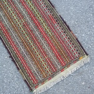 May include: A long, narrow rug with a geometric pattern in shades of red, green, brown, and white. The rug has a fringe on both ends.