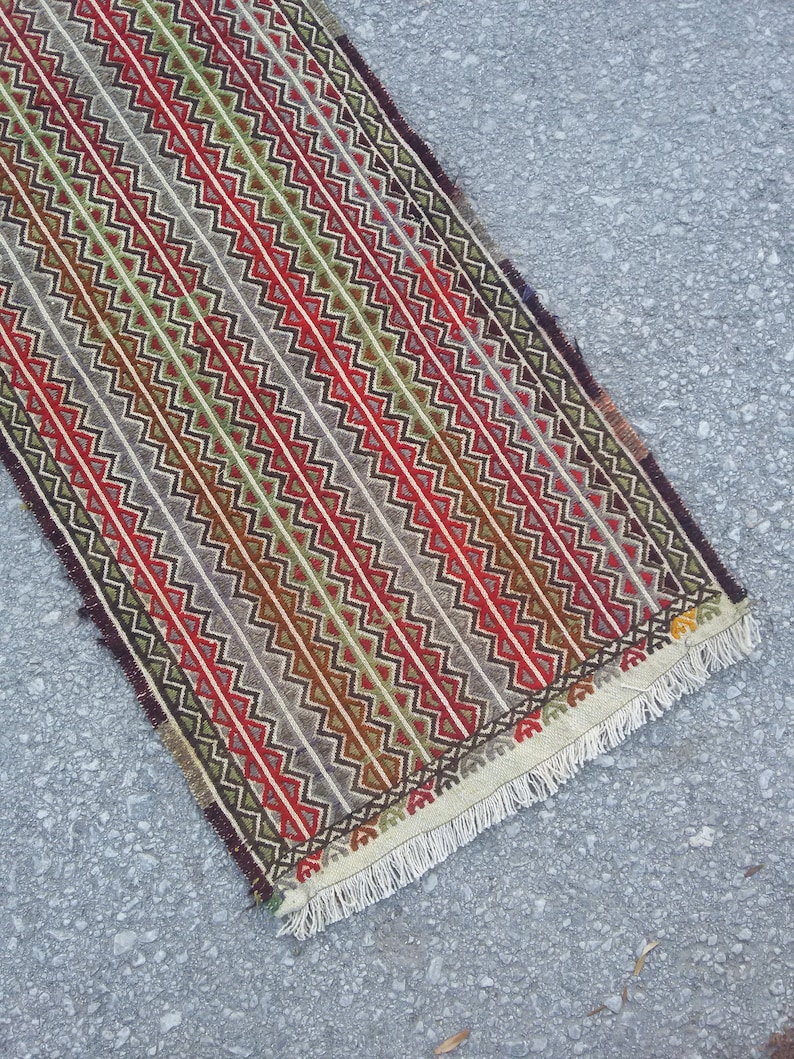 May include: A long, narrow rug with a geometric pattern in shades of red, green, brown, and white. The rug has a fringe on both ends.