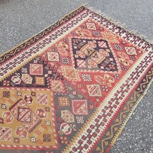 May include: A traditional Turkish rug with a geometric pattern in shades of red, brown, beige, and white. The rug has a fringe border and is woven with intricate details.
