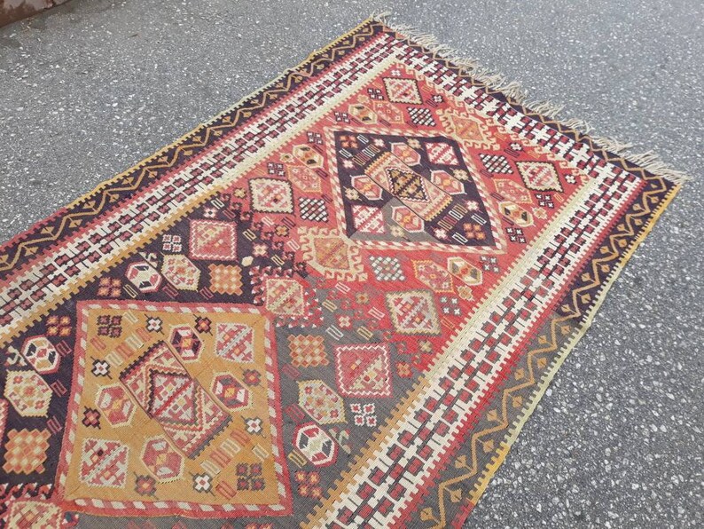 May include: A traditional Turkish rug with a geometric pattern in shades of red, brown, beige, and white. The rug has a fringe border and is woven with intricate details.