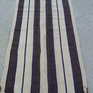 May include: A long, narrow, cream-colored rug with alternating black and purple stripes. The rug is woven with a simple, rustic design.