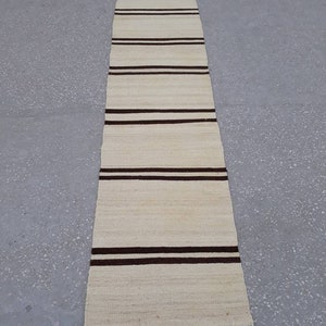 May include: A long, narrow, off-white rug with brown stripes. The rug has a fringed edge.