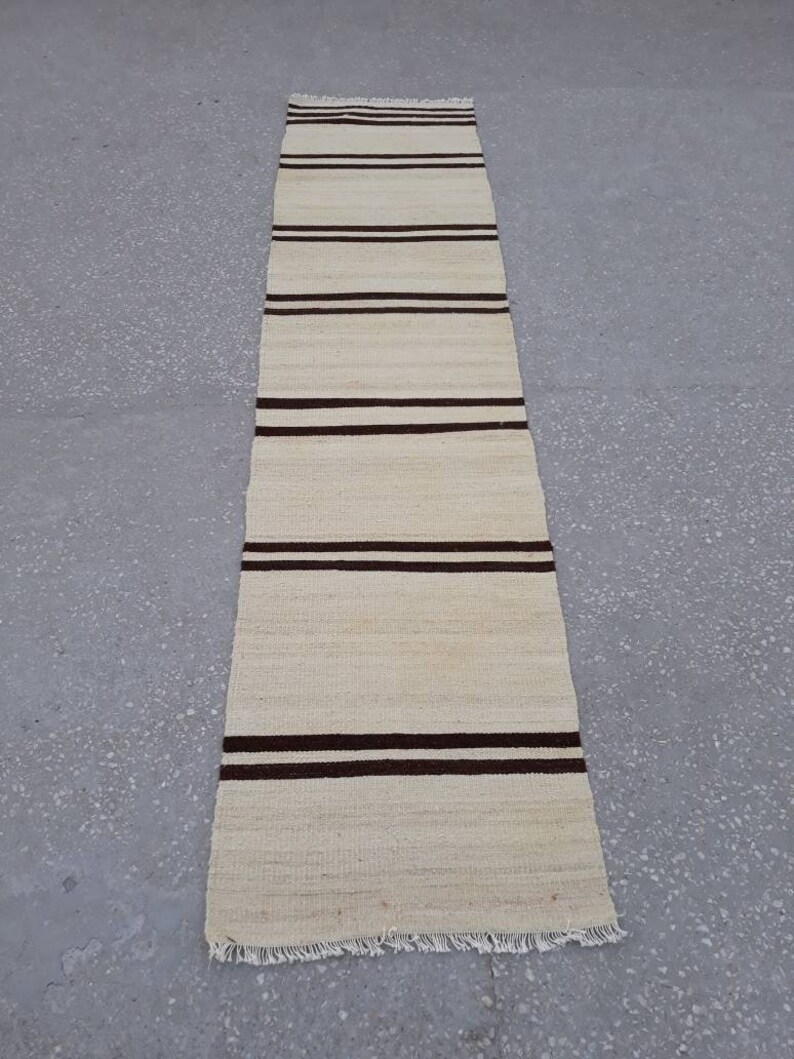 May include: A long, narrow, off-white rug with brown stripes. The rug has a fringed edge.