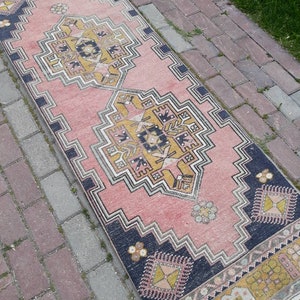 May include: A pink and yellow patterned runner rug with a geometric design. The rug has a dark blue border and is laid out on a brick walkway.