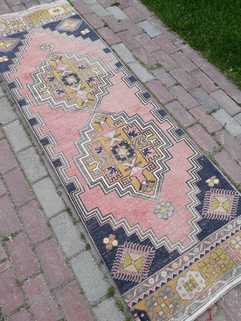 May include: A pink and yellow patterned runner rug with a geometric design. The rug has a dark blue border and is laid out on a brick walkway.