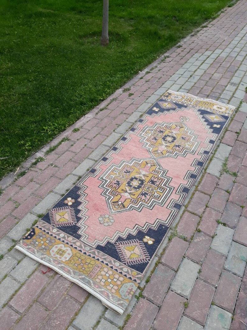 May include: A pink and blue patterned runner rug with a geometric design. The rug is laid out on a brick walkway.