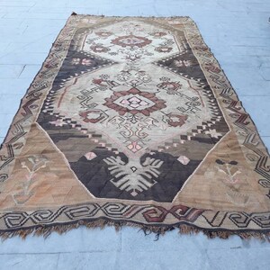 May include: A vintage Turkish rug with a brown, beige, and pink geometric pattern. The rug has a fringed edge and is made of wool.