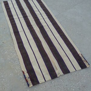 May include: A long, narrow, striped rug with alternating white and dark brown stripes. The rug is woven with a simple, rustic design and has a frayed edge.